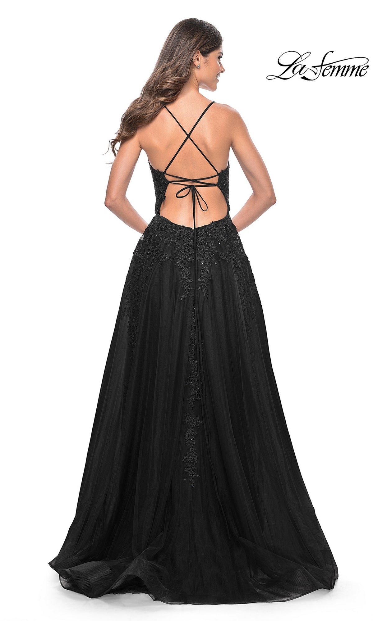 A woman stands facing away, wearing the La Femme Long Lace A-Line Prom Dress 32303—a floor-length black gown with lace details, an open back, and crisscross straps.